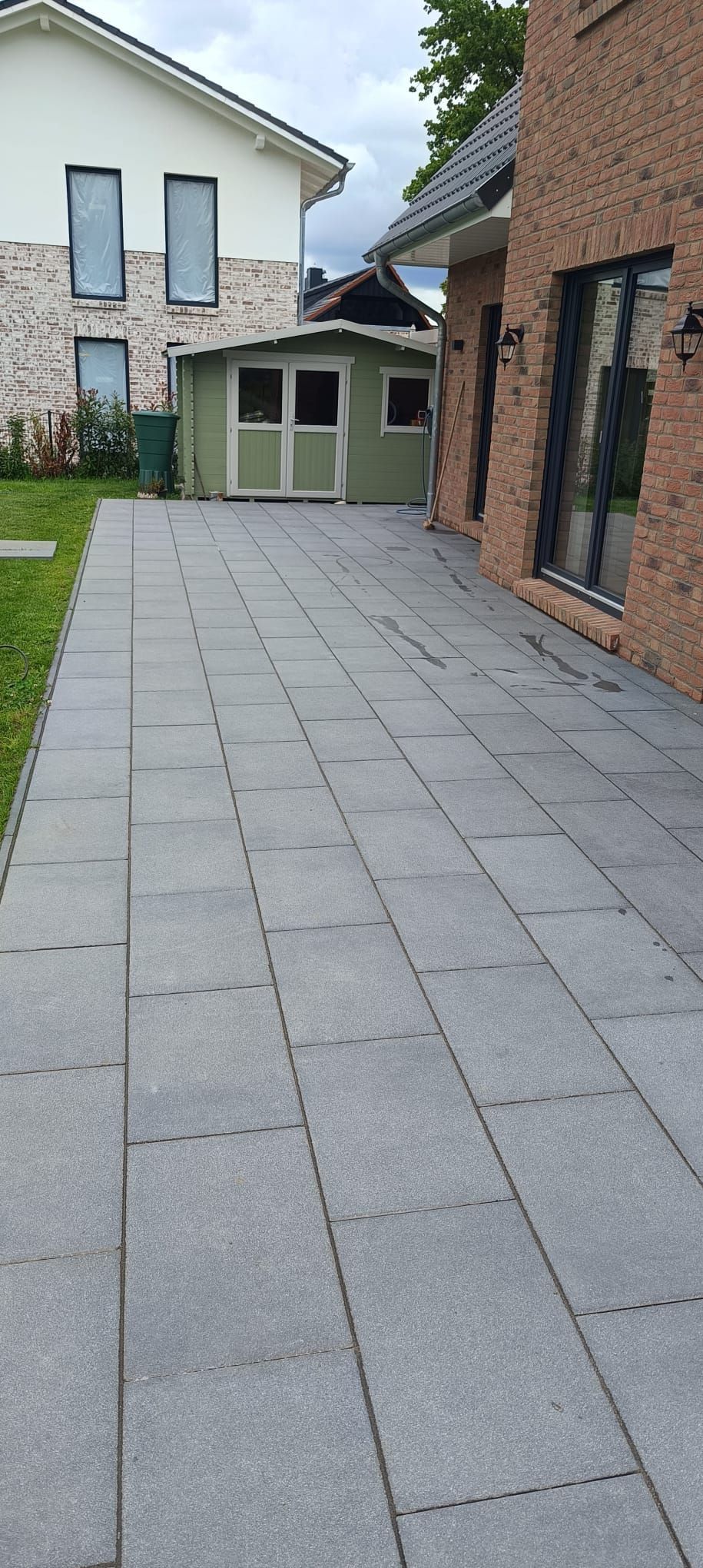 Ein gepflasterter Fußweg führt vorbei an einem Backsteingebäude und einem grasbewachsenen Hof zu einem grünen Schuppen und einem weißen Haus.