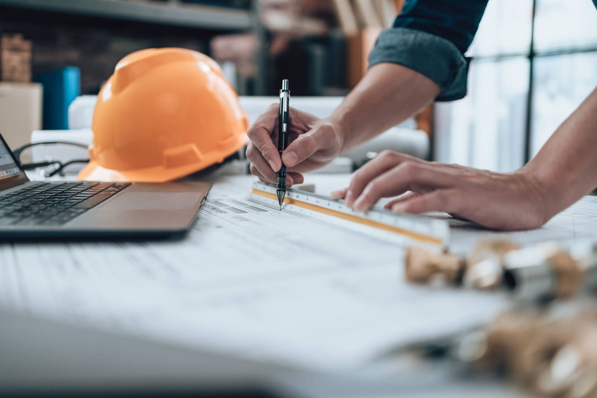 Un trabajador de la construcción está trabajando en un plano con una computadora portátil y un casco en el fondo.