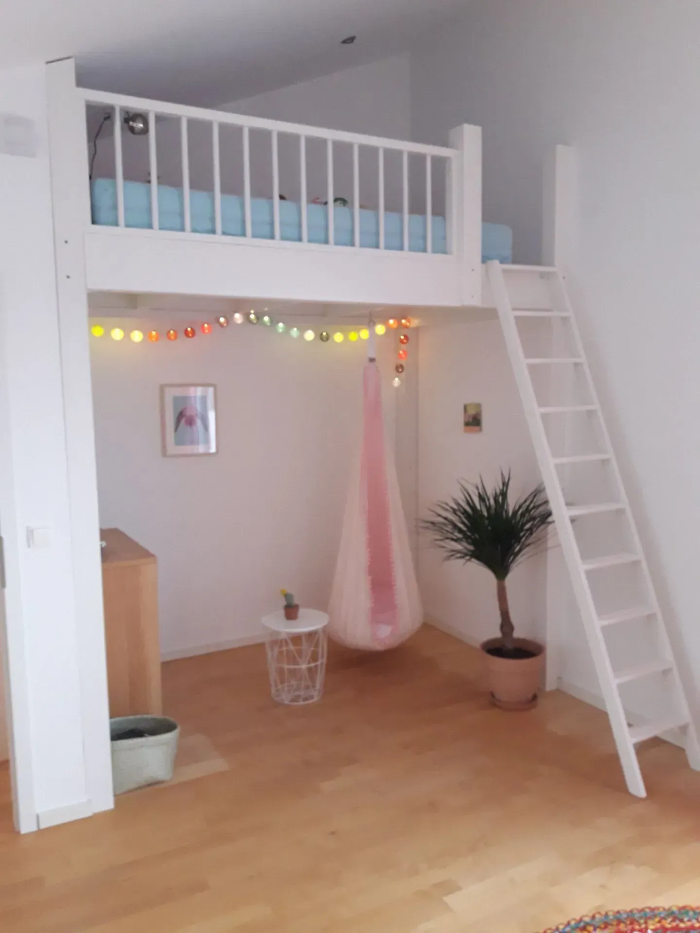Weißes Hochbett mit Leiter, Lichterkette, rosa Hängesessel und Topfpflanze in einem hellen Zimmer.