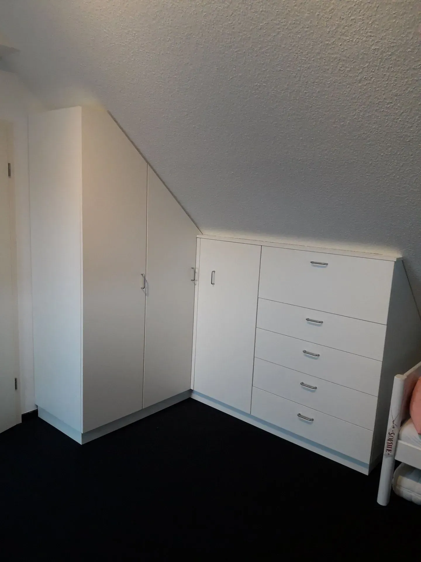 Weiße Einbauschränke in einem Zimmer mit Dachschräge und schwarzem Teppichboden.