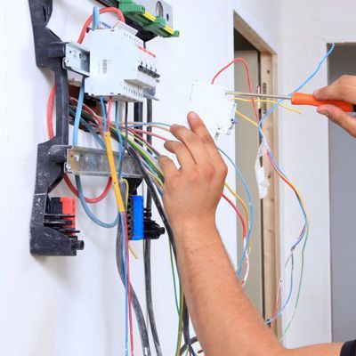 Un electricista utiliza un destornillador para conectar componentes a un panel de disyuntores abierto en una pared blanca.