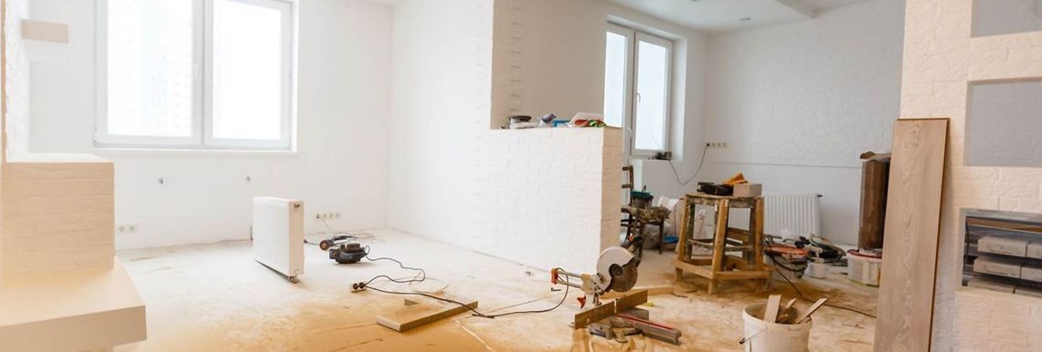 Una habitación luminosa y sin terminar durante la reforma de una vivienda, con paredes blancas al descubierto, escombros de construcción en el suelo y una escalera.