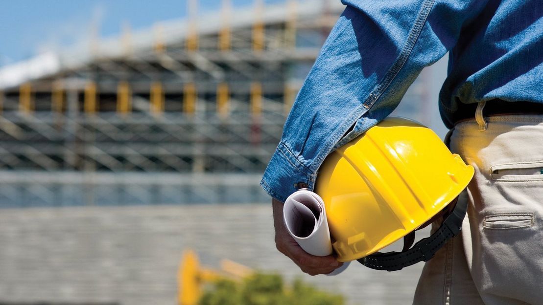 Una persona con una camisa vaquera sostiene un casco amarillo y unos planos enrollados frente a una obra en construcción borrosa.