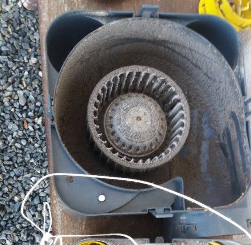 Vue de dessus d'un ventilateur gris à hélice circulaire, posé sur du gravier.