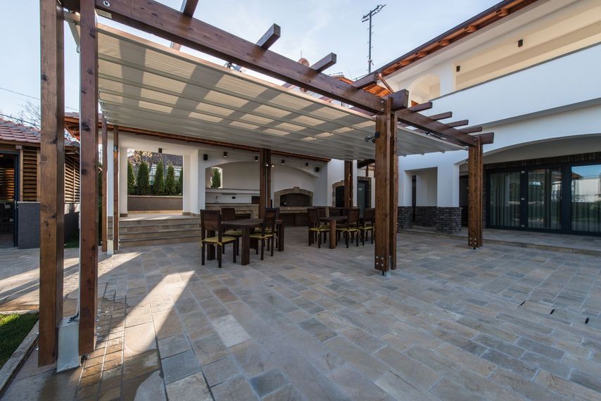 Patio moderne avec pergola, tables et chaises, sol en pierre et un bâtiment avec portes vitrées.