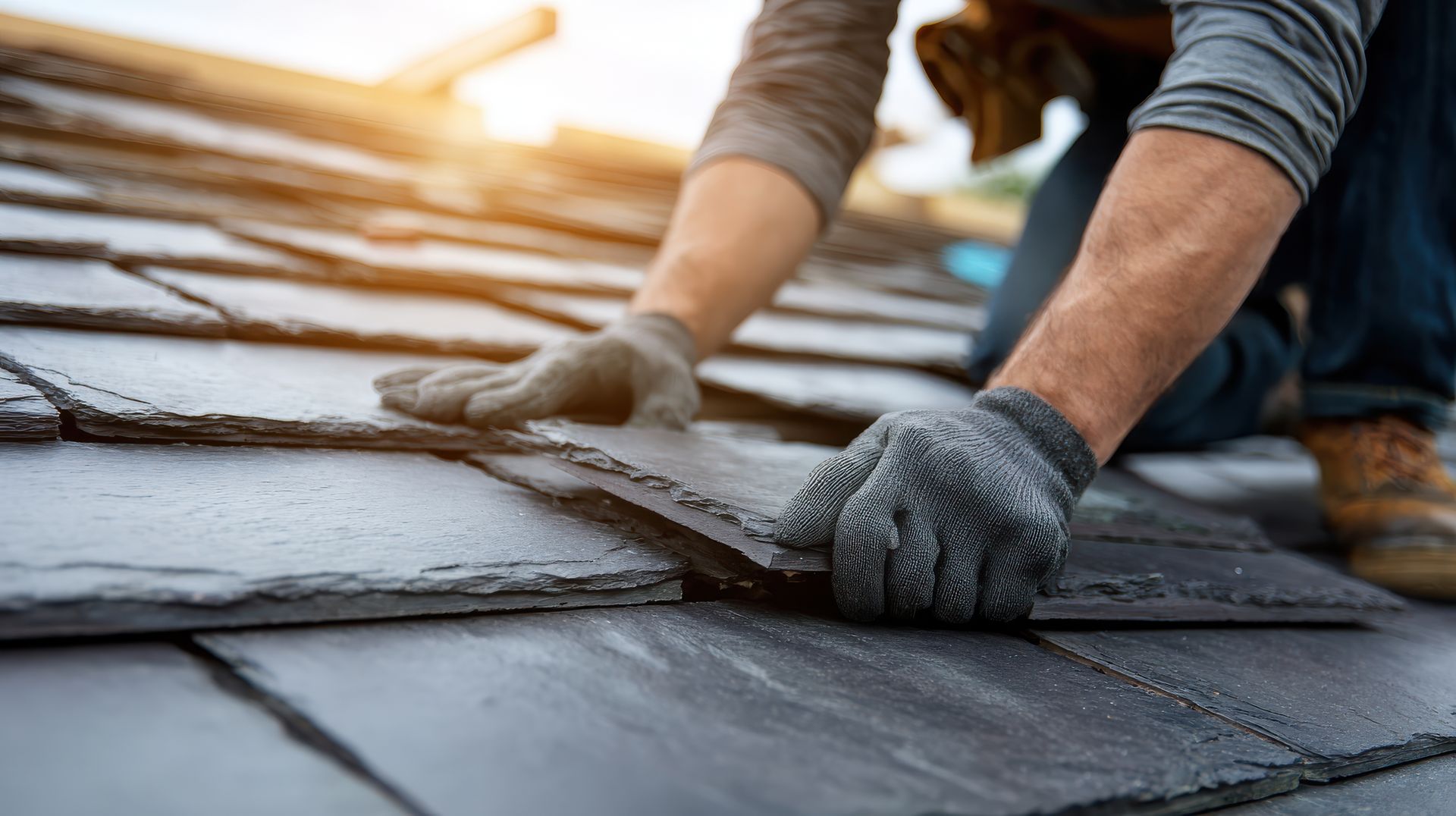 Un ouvrier portant des gants de protection installe des tuiles d'ardoise foncées sur un toit par une journée ensoleillée.