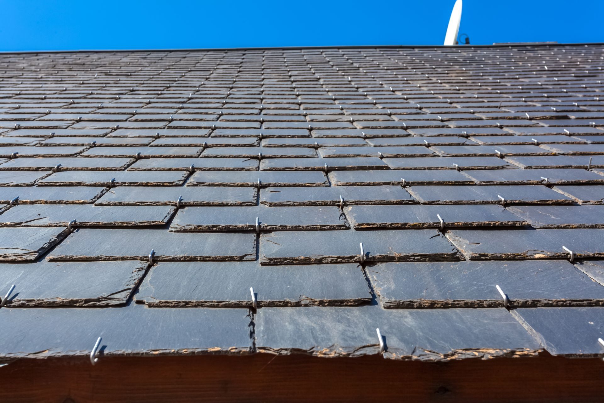 Gros plan sur un toit d'ardoise sombre se détachant sur un ciel bleu vif, montrant les fixations métalliques visibles sur chaque tuile.