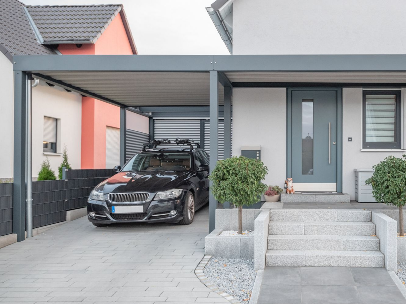 Un carport moderne gris abritant une voiture noire, attenant à une maison aux portes grises, à l'escalier clair et aux deux petits arbres ronds.