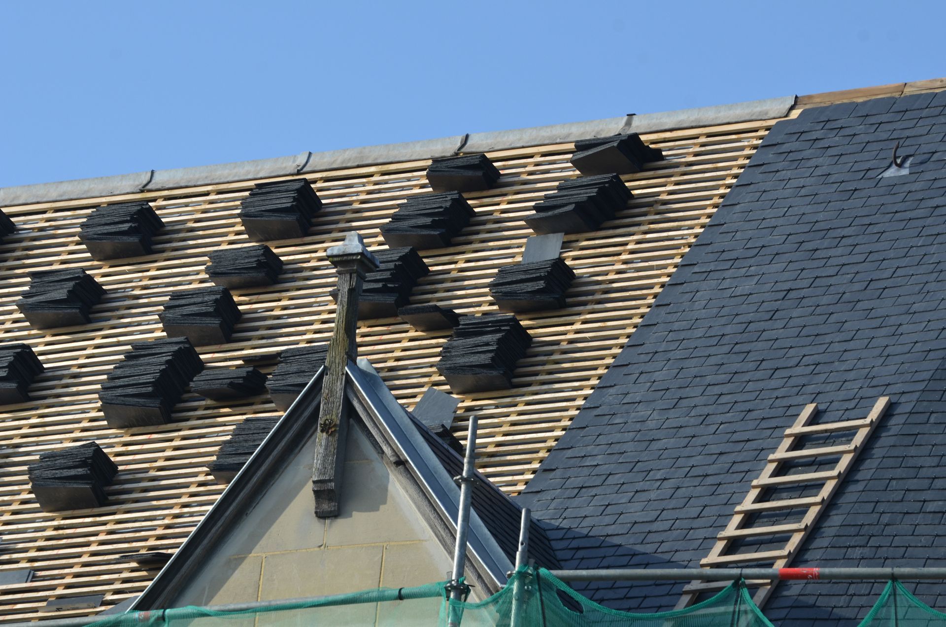 Un toit partiellement refait à neuf, avec des piles de tuiles d'ardoise noire posées sur les liteaux en bois et une échelle sur la droite.