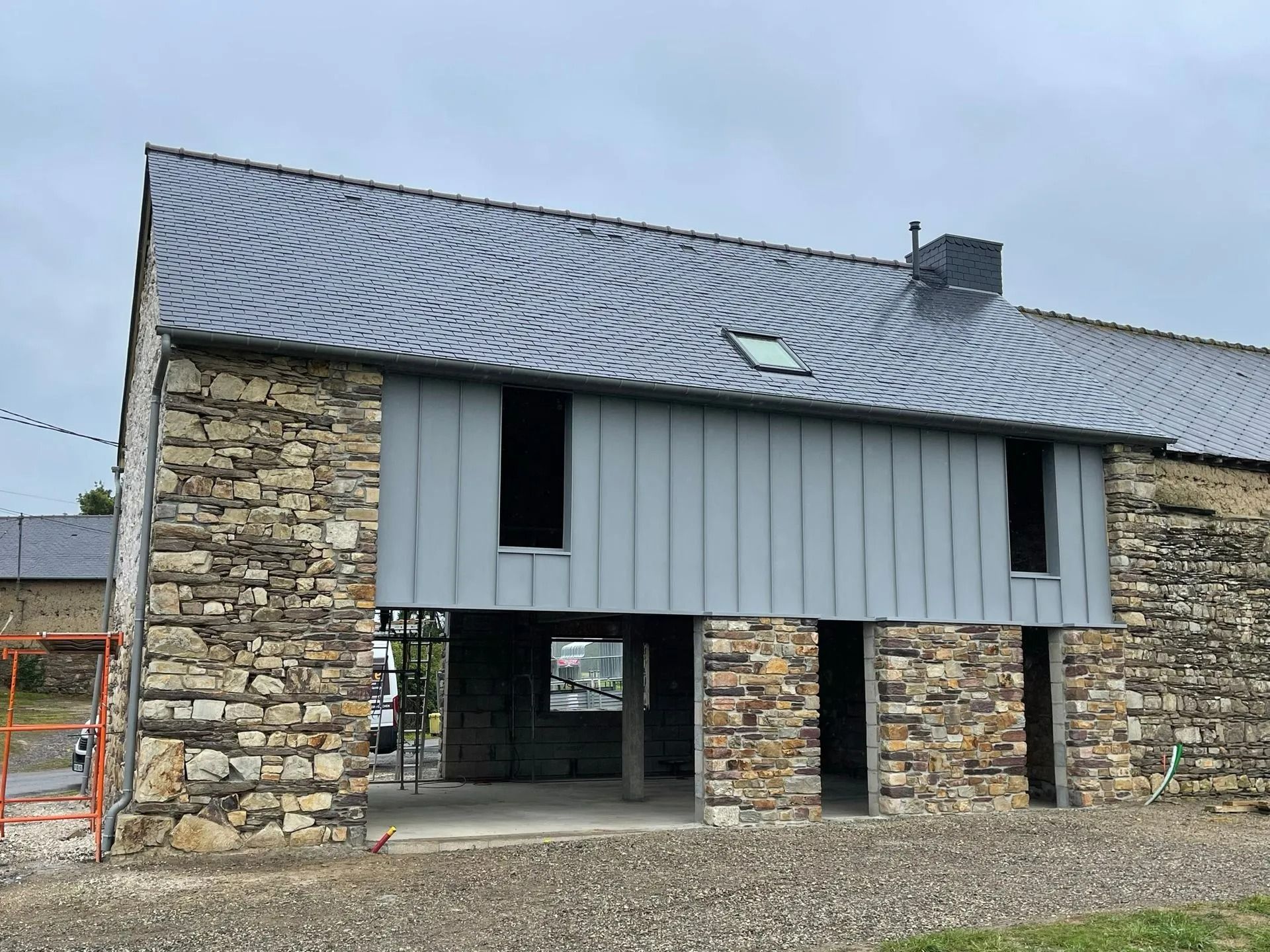 Un bâtiment en pierre rénové, en cours de construction, avec un bardage métallique gris, un toit en ardoise et des portes ouvertes.