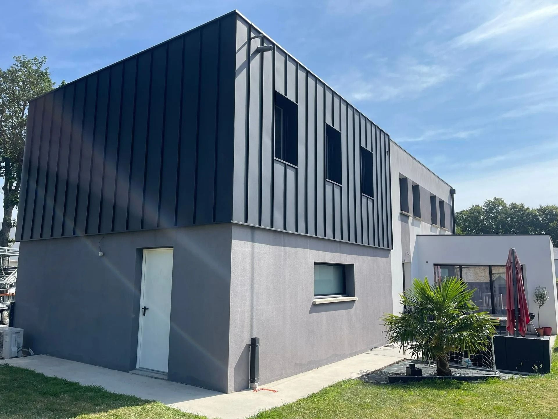 Une maison moderne à deux étages, avec un niveau supérieur en métal gris foncé et un niveau inférieur en stuc gris clair, sous un ciel bleu azur.
