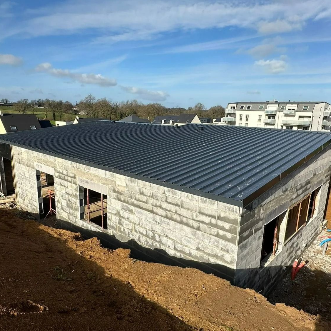 Un bâtiment inachevé en parpaings gris, avec un toit en tôle ondulée sombre, se dresse sur un chantier sous un ciel bleu.