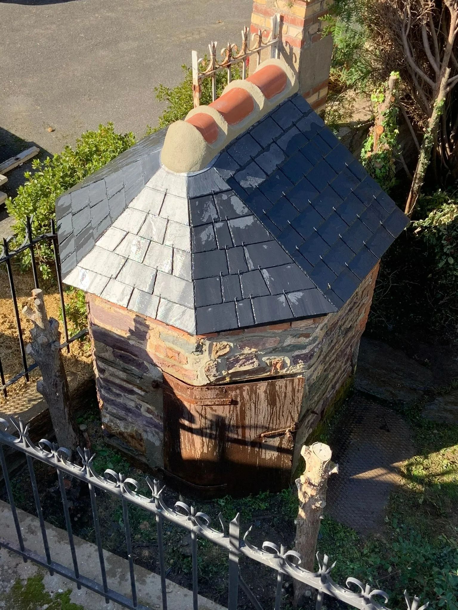 Une petite structure rectangulaire en pierre, avec un toit à quatre pans en ardoise et une crête en terre cuite, située dans un jardin.