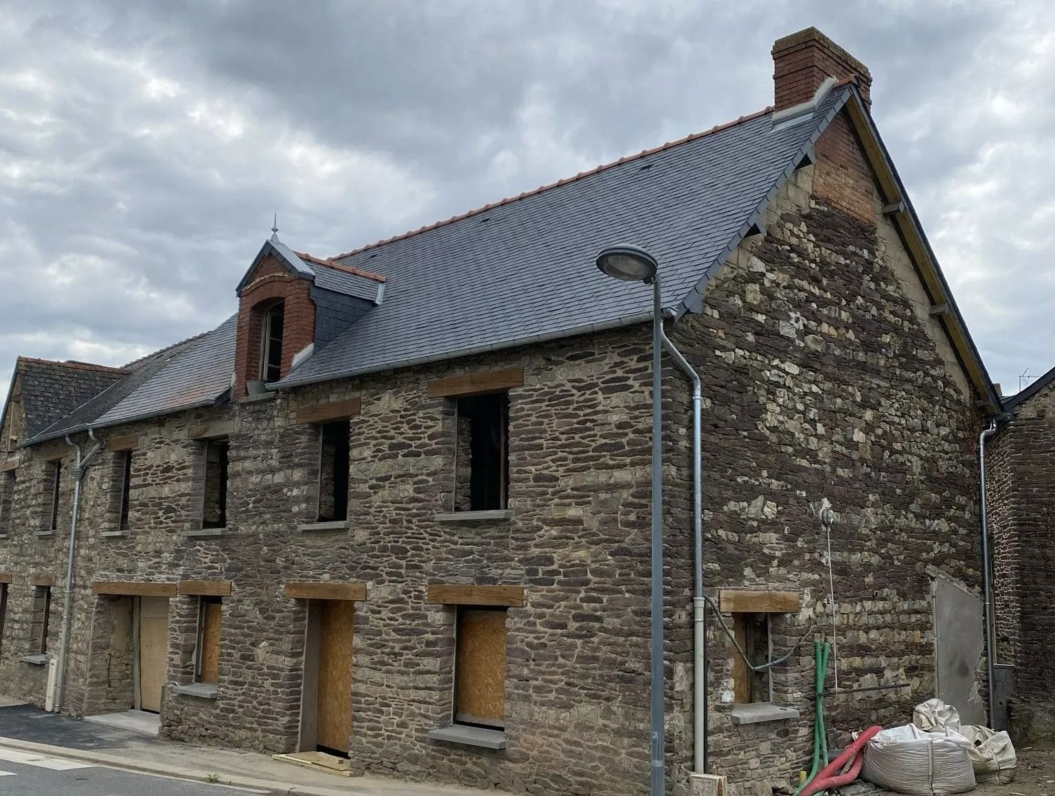 Un immeuble en pierre à plusieurs étages en cours de rénovation, avec un nouveau toit gris, des fenêtres condamnées et une cheminée se détachant sur un ciel nuageux.
