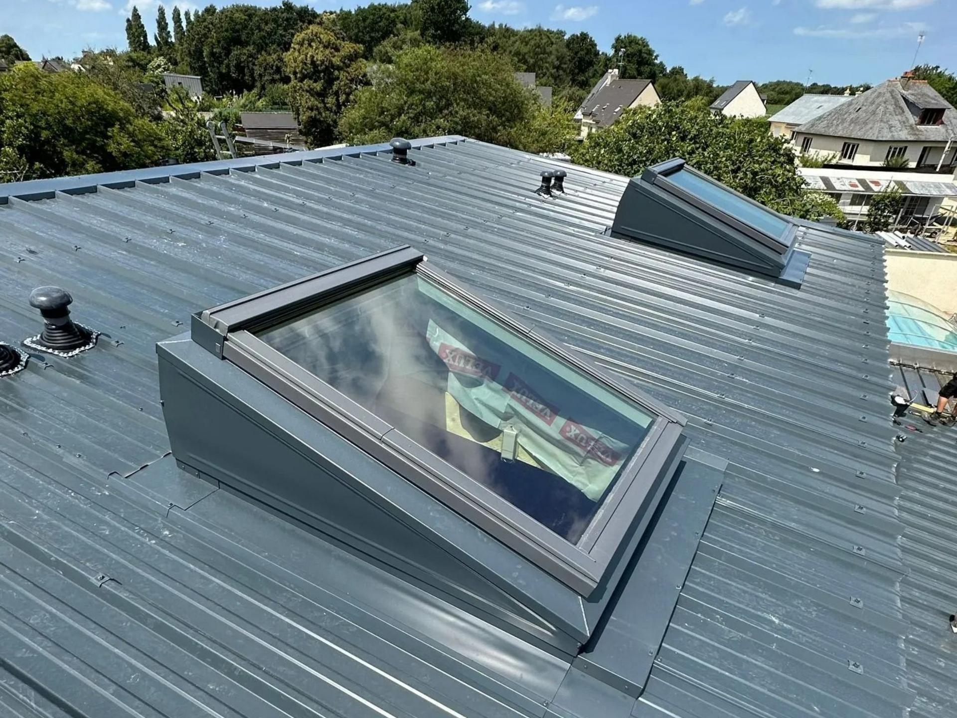 Deux puits de lumière installés sur un toit en tôle ondulée grise, sous un ciel bleu clair et lumineux.