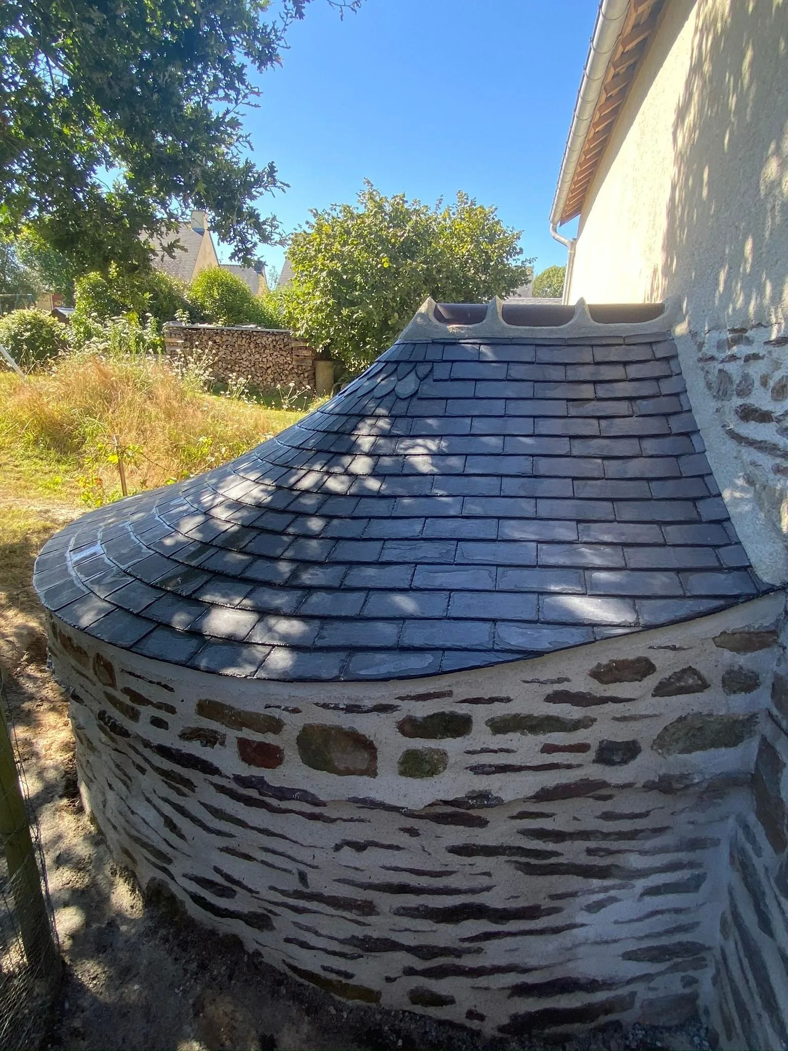 Un mur de pierre incurvé soutient un toit conique en bardeaux sombres qui se prolonge sur le côté d'un bâtiment sous un ciel d'un bleu éclatant.