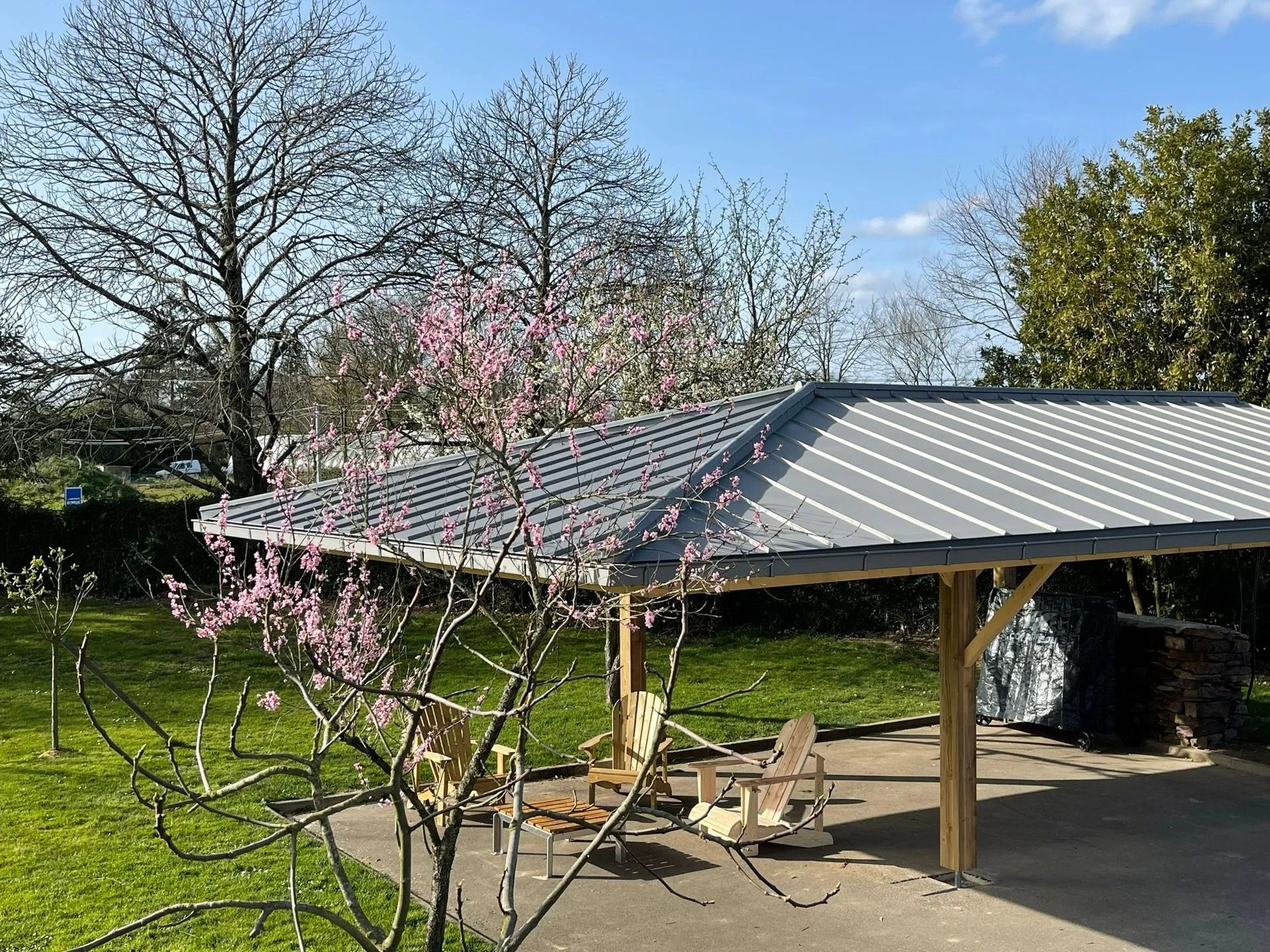 Une terrasse couverte avec des poteaux en bois clair et un toit en métal, agrémentée de chaises d'extérieur par une journée ensoleillée, avec des arbres en fleurs à proximité.