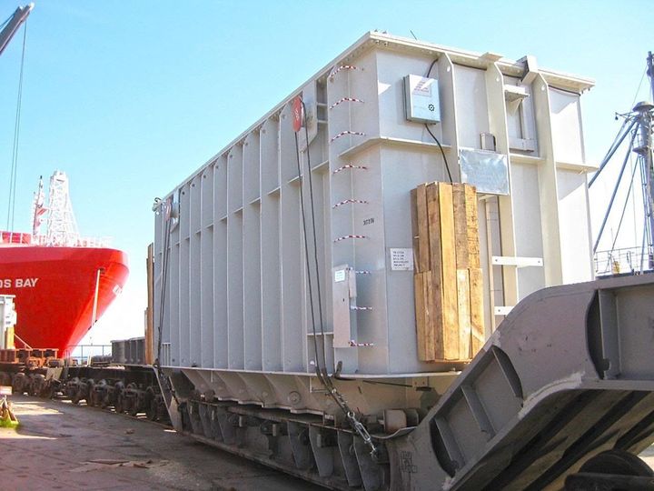 Large gray industrial cargo container on a flatbed railcar in a port beside a red ship