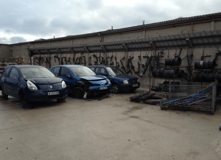 Trois voitures endommagées dans une casse, garées près de pièces détachées et de pneus.