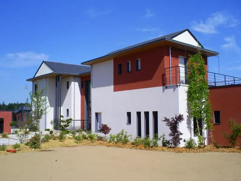 Bâtiment moderne de deux étages aux murs blancs et brun rougeâtre, au toit sombre et doté d'un petit balcon se détachant sur un ciel bleu.