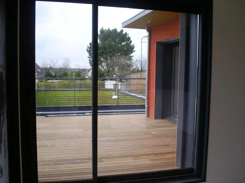 Vue par une fenêtre sur une terrasse en bois, une pelouse et une maison au mur orange.