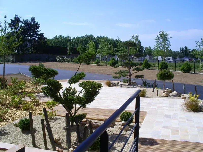 Vue d'un espace extérieur aménagé avec divers arbustes taillés, des arbres et des allées de pierre sous un ciel d'un bleu éclatant.