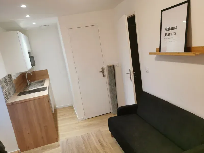 Petit studio avec une kitchenette et un canapé noir, des murs blancs.