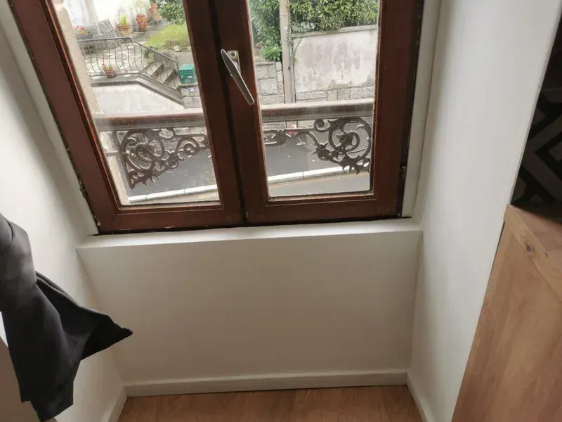 Fenêtre dans une pièce laissant apparaître, à travers la vitre, une balustrade de balcon noire décorative. Murs blancs et boiseries.