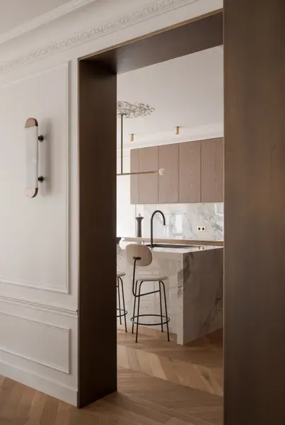 Vue intérieure : Coin cuisine visible à travers une porte, avec un parquet clair, des murs aux tons neutres et un îlot en marbre.