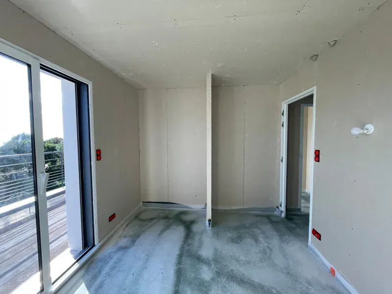 Pièce intérieure en construction avec des murs clairs, une porte et une grande fenêtre donnant sur un balcon.