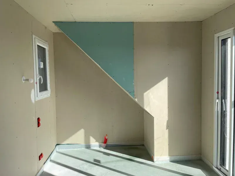 Pièce intérieure aux murs bruts et aux accents bleu clair. Sol, fenêtre et porte baignés de soleil.