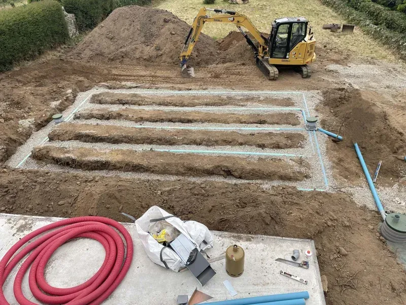 Une excavatrice creuse des tranchées pour un système de drainage ; des monticules de terre et des tuyaux sont visibles.