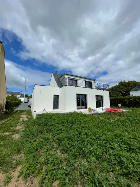 Maison moderne blanche aux fenêtres noires sous un ciel bleu nuageux. Pelouse verte au premier plan.