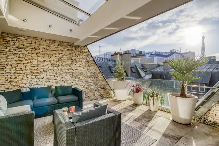 Terrasse sur le toit avec un canapé bleu, des plantes en pot, un mur en pierre, un toit en verre et une vue sur la tour Eiffel.