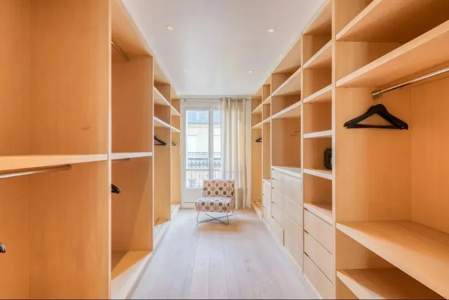 Dressing avec des étagères en bois clair, des tiroirs et une tringle à vêtements avec une chaise près d'une fenêtre.