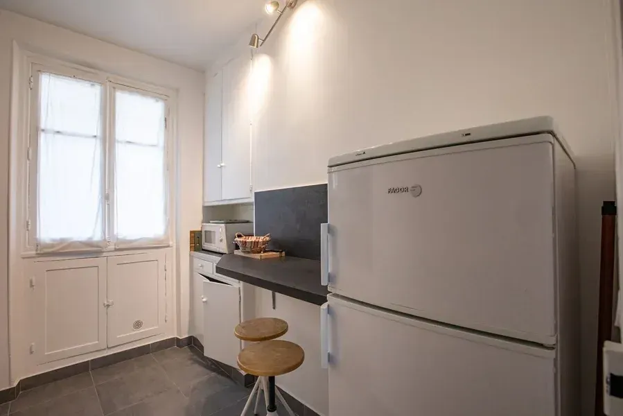 Petite cuisine avec électroménager blanc, des placards blancs et un plan de travail noir. Une fenêtre se trouve dans un coin.