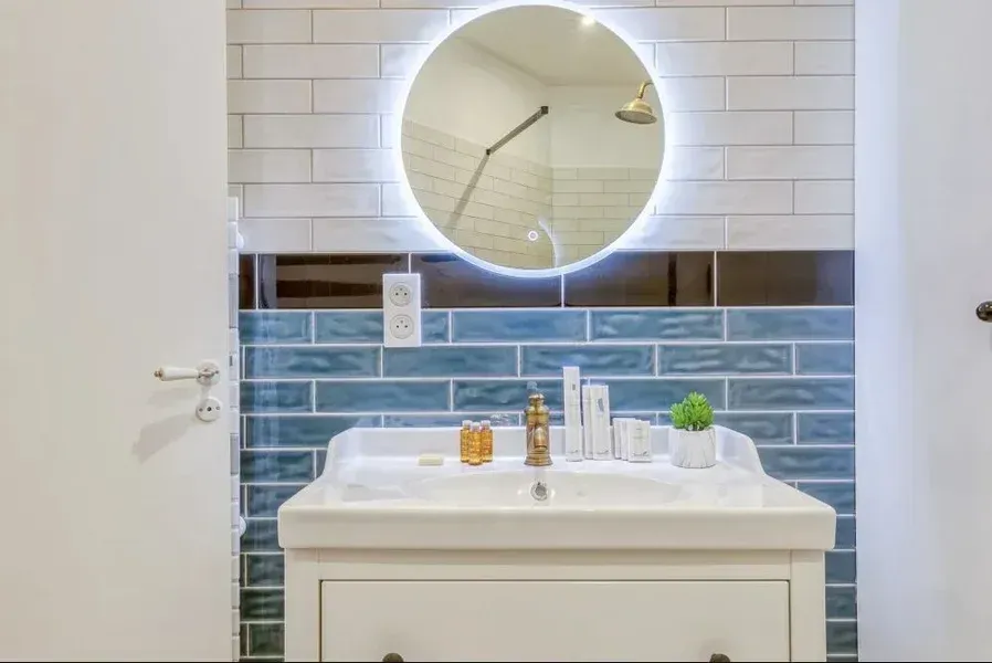 Salle de bains avec un lavabo blanc, des murs carrelés bleu et blanc et un miroir rond à bord lumineux.