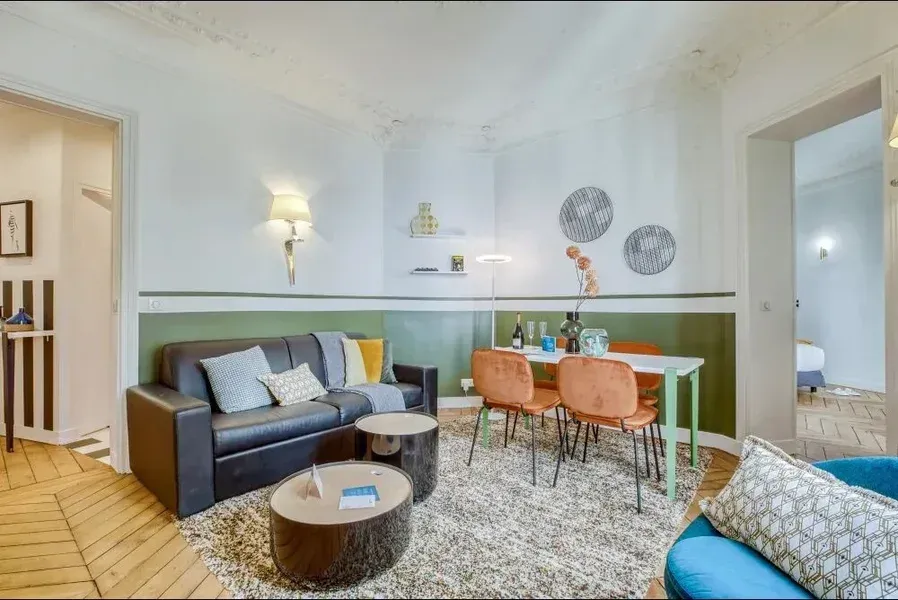 Salon avec un canapé noir, des chaises orange, un tapis et une petite table à manger.