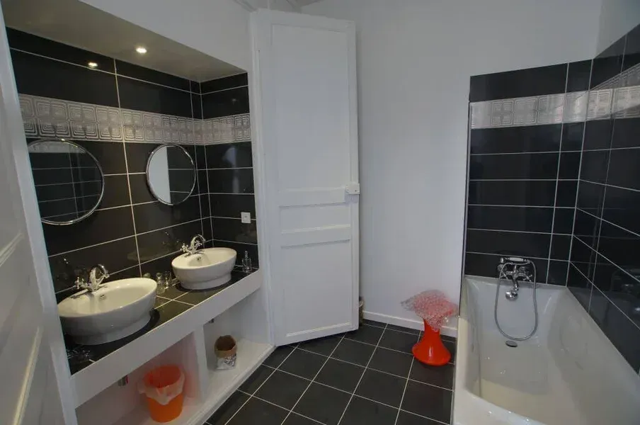 Salle de bains avec un carrelage noir, des lavabos blancs et une baignoire. Une porte blanche est placée au centre.