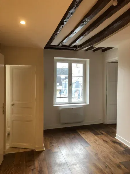 Pièce intérieure avec des poutres apparentes, des murs blancs, une fenêtre, un parquet et une porte ouverte.