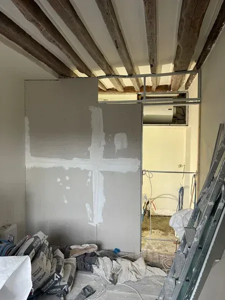 Rénovation intérieure : pose de plaques de plâtre dans une pièce avec des poutres apparentes au plafond et une porte.