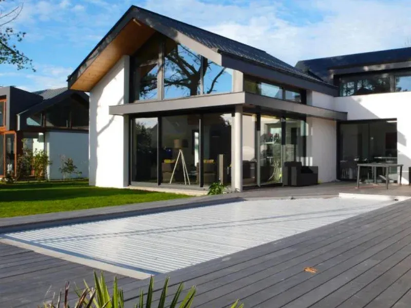 Maison blanche moderne avec des boiseries foncées et de grandes fenêtres, donnant sur un patio gris.