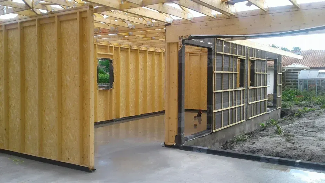 Structure en bois d'un bâtiment en construction, avec un côté ouvert. L'intérieur révèle la charpente et une fenêtre.