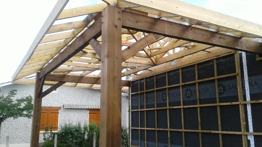Pergola en bois en construction ; poutres et chevrons apparents, mur sombre.
