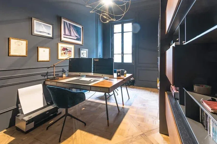 Bureau à domicile avec des murs bleu foncé, un bureau avec deux écrans et des œuvres d'art.