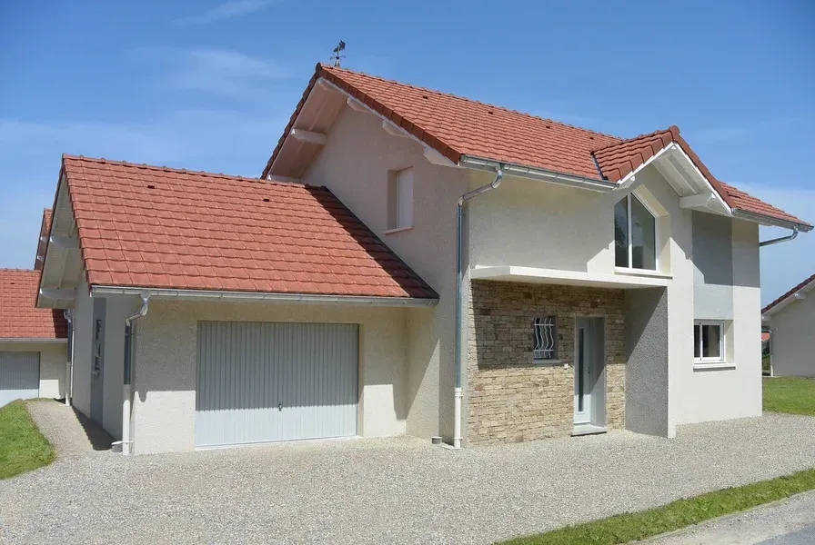 Maison à deux étages avec un toit rouge et un garage ; Une allée de gravier, un ciel bleu.