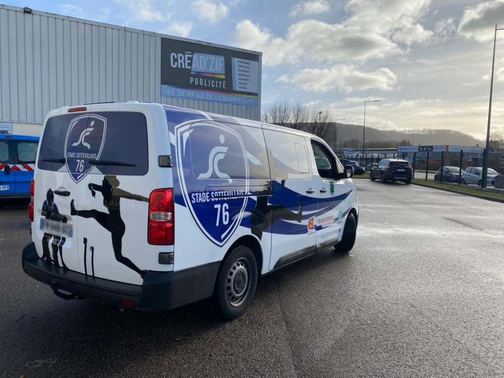 Fourgon de sport blanc et bleu garé devant un entrepôt, sur un parking détrempé, avec le numéro 76 sur le côté