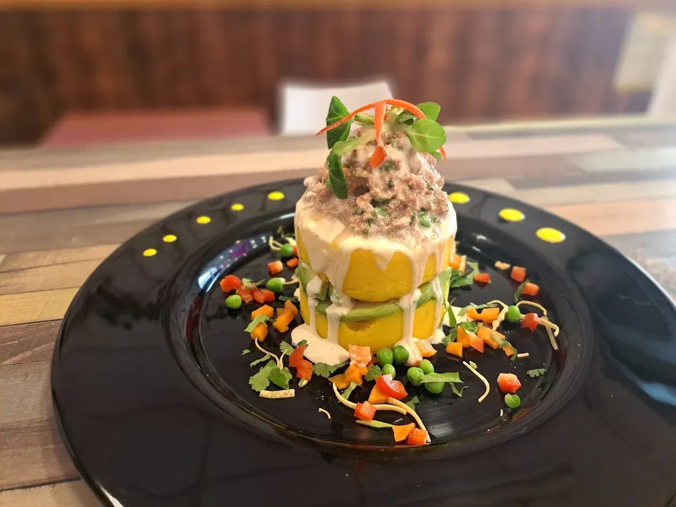 Torre de comida en un plato negro, con capas de amarillo, verde y blanco. Decorada con verduras y hierbas.