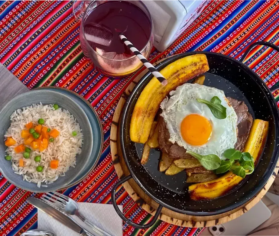 Plato de bistec y huevo frito con plátanos, papas y arroz. Bebida morada. Mantel colorido.