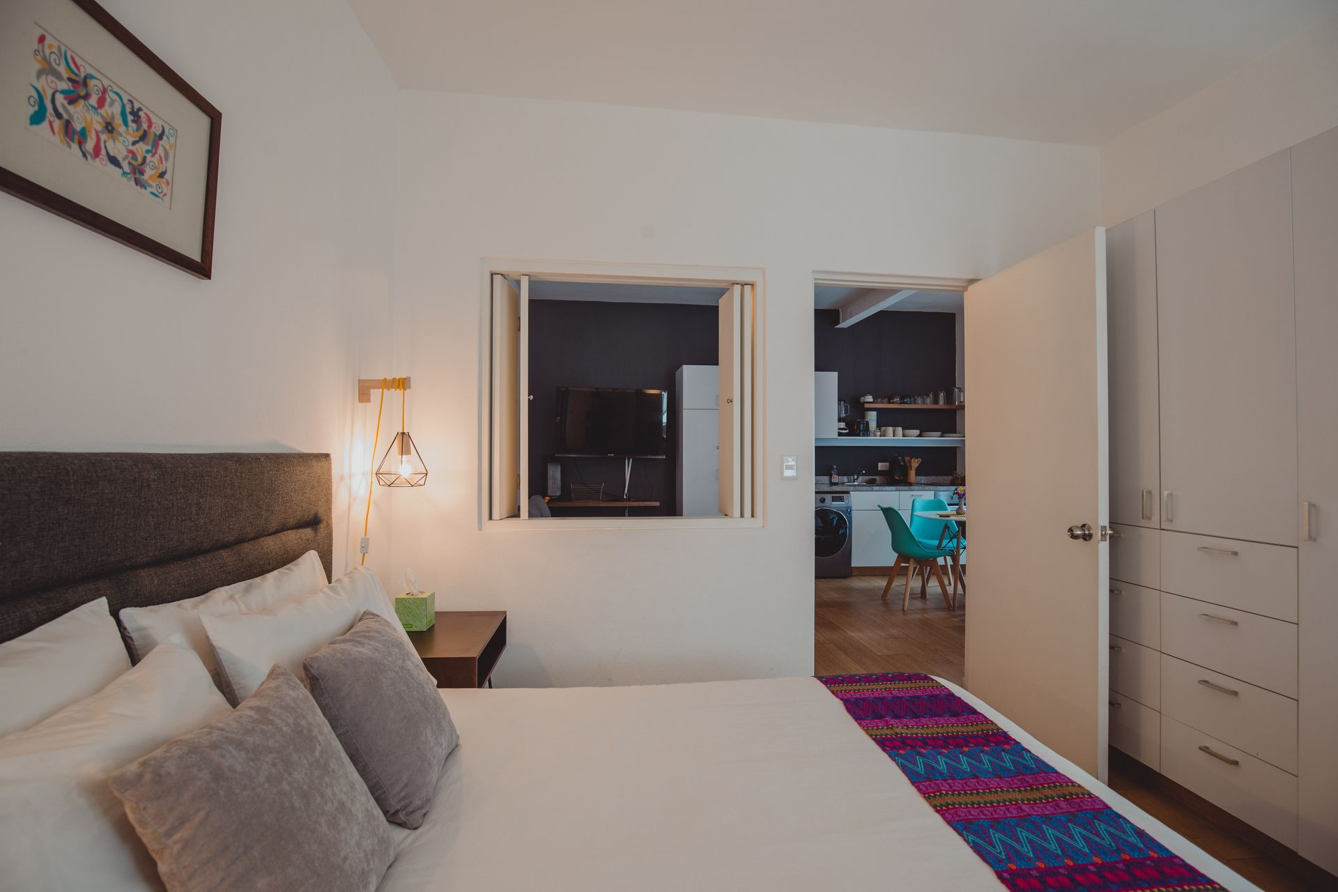A bedroom with a large bed and a window looking into the kitchen.
